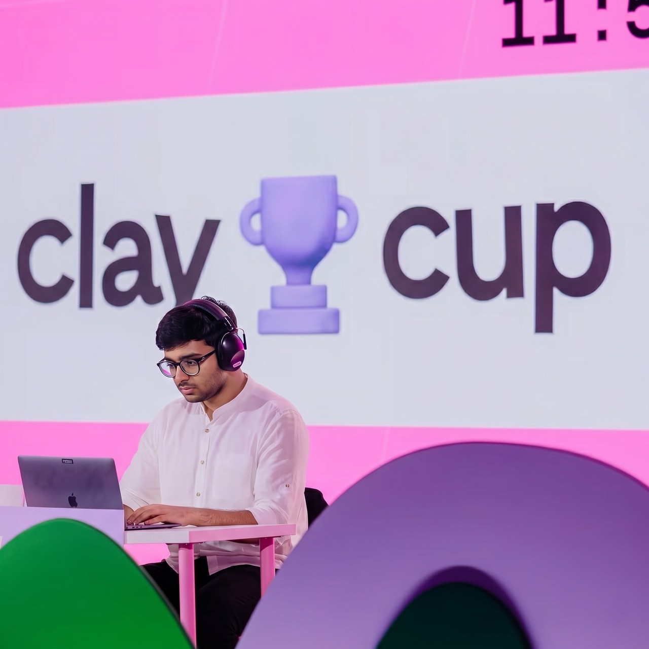 Man in white shirt with headphones working on a laptop under a pink Clay Cup sign with a trophy icon.